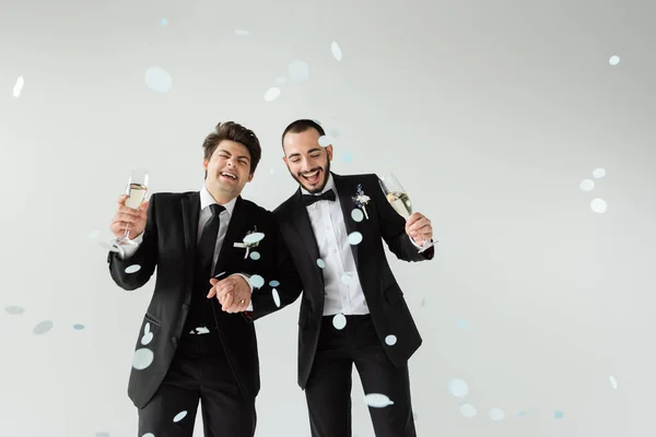 Excited homosexual grooms in elegant formal wear holding hands and glasses of champagne while standing under falling confetti during wedding on grey background — Stock Photo