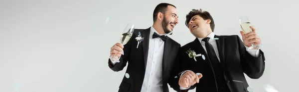Excited homosexual grooms in classis attire holding hands and glasses of champagne under falling confetti during wedding celebration on grey background, banner — Stock Photo