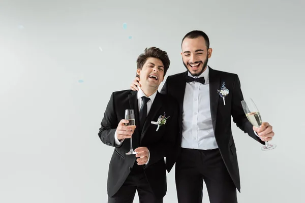 Excited same sex grooms in formal wear with floral boutonnieres holding champagne while celebrating wedding under falling confetti on grey background — Stock Photo