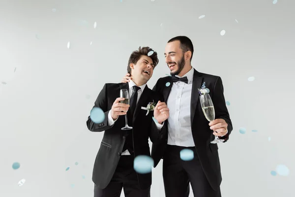 Alegre homossexual noivo no elegante terno abraçando jovem namorado com copo de champanhe enquanto celebrando casamento sob queda confetti no fundo cinza — Fotografia de Stock