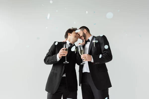 Cheerful homosexual grooms in suits touching each other while holding glasses of champagne and celebrating marriage under falling confetti on grey background — Stock Photo