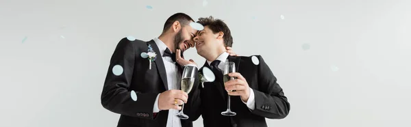 Unbekümmerte gleichgeschlechtliche Bräutigame in Anzügen, die sich berühren und Champagner halten, während sie ihre Hochzeit unter fallendem Festkonfetti auf grauem Hintergrund feiern, Banner — Stockfoto
