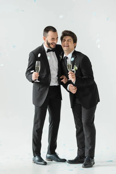 Full length of overjoyed same sex grooms in suits holding champagne and having fun under falling festive confetti during their wedding day on grey background — Stock Photo