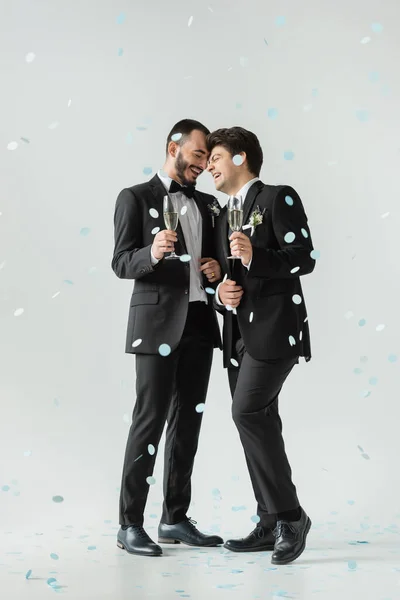 Full length of positive same sex couple in classic suits holding glasses of champagne while celebrating wedding under falling confetti on grey background — Stock Photo