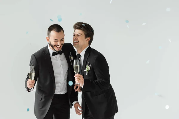 Веселий гей наречений тримає шампанське біля елегантного хлопця в класичному костюмі, стоячи під падінням конфетті під час весілля на сірому фоні — стокове фото