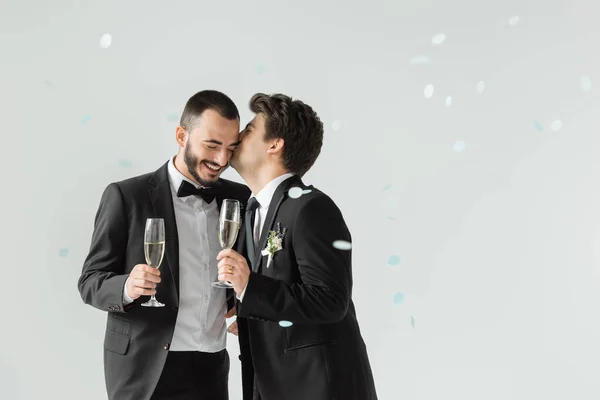 Jovem noivo homossexual em desgaste formal beijando namorado barbudo segurando champanhe sob queda confetti no fundo cinza — Fotografia de Stock