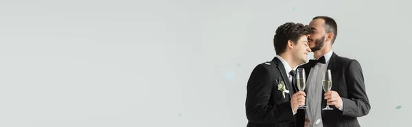 Giovane uomo omosessuale in tuta che tiene champagne e bacia fidanzato sorridente con champagne sotto coriandoli durante la cerimonia nuziale su sfondo grigio, banner — Foto stock