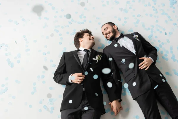 Top view of positive same sex grooms in formal wear looking at each other and holding hands while lying under falling confetti on grey background — Stock Photo