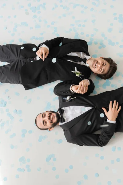 Vue du dessus des mariés gays gais en classe tenue tenant la main et regardant la caméra tout en étant couché sur des confettis bleus pendant le mariage sur fond gris — Photo de stock