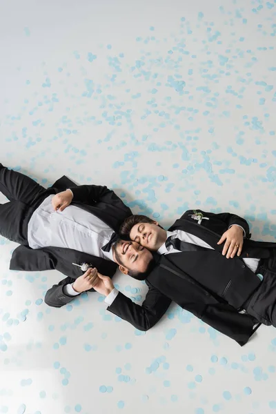 Вид зверху на молодих і усміхнених гомосексуальних наречених із закритими очима в костюмах, тримаючись за руки, лежачи разом на святковому конфеті на сірому фоні — стокове фото
