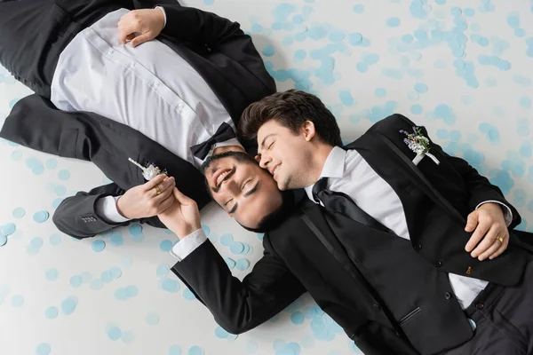 Top view of smiling gay groom touching hand of bearded boyfriend in classic suit lying together on festive confetti during wedding celebration on grey background — Stock Photo