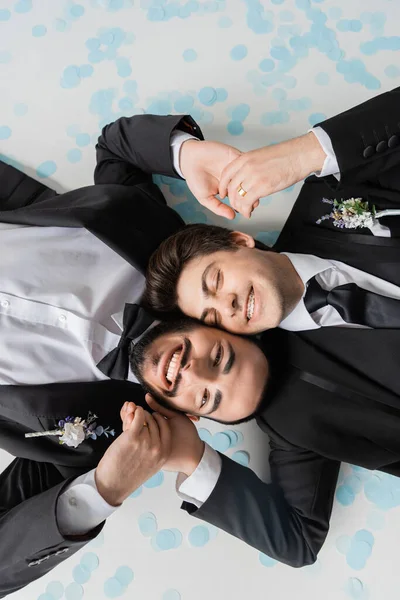 Top view of cheerful same sex couple in suits holding hands while lying together on confetti and smiling during wedding celebration on grey background — Stock Photo