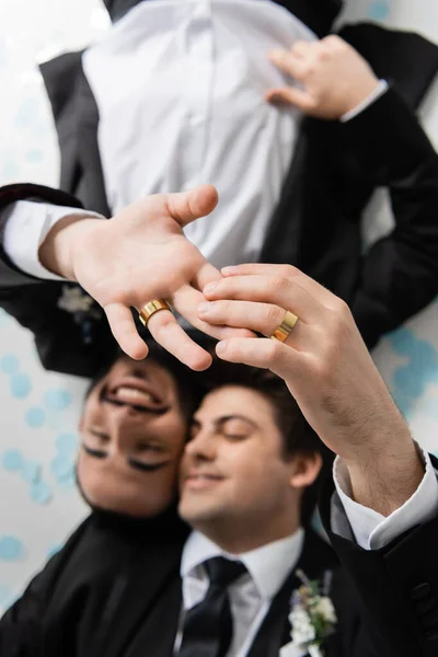 Draufsicht auf verschwommene gleichgeschlechtliche Bräutigame in Anzügen, die sich in Eheringen gegenseitig die Finger berühren, während sie auf festlichem Konfetti auf grauem Hintergrund liegen — Stockfoto