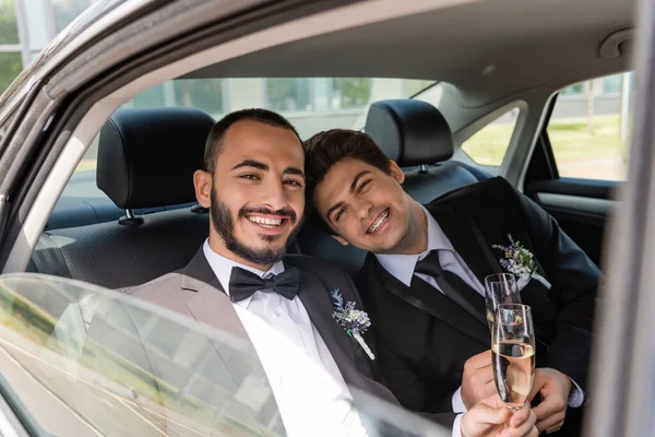 Young and positive newlyweds in formal wear with floral boutonnieres holding glasses of champagne and looking at camera from car window while sitting on backseat — Stock Photo