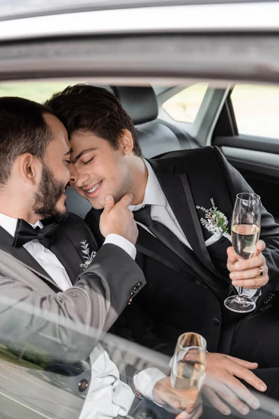 Веселый гей-жених в классическом костюме с бутоньерным трогательным подбородком молодого парня в брекетах и с бокалом шампанского в руках, сидя на заднем сидении автомобиля — стоковое фото
