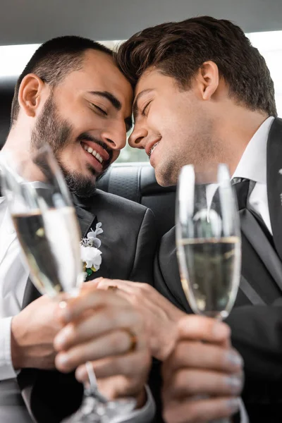 Portrait of overjoyed gay newlyweds in classic attire holding blurred hands and glasses of champagne during honeymoon road trip in car — Stock Photo