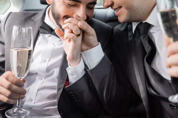 Smiling and bearded gay man with wedding ring on hand kissing hand of boyfriend in braces and holding blurred champagne while sitting in car during honeymoon — Stock Photo