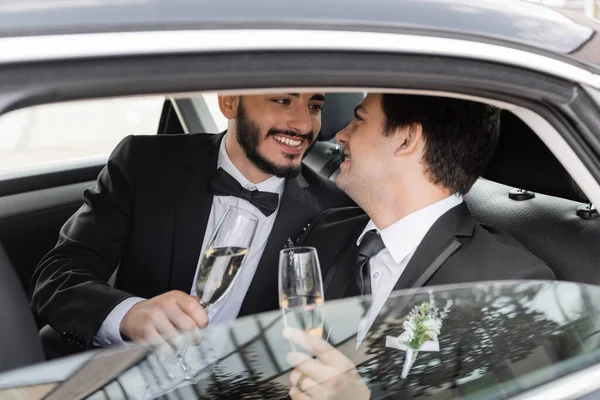 Positive and bearded homosexual groom in elegant suit holding glass of champagne and looking at young boyfriend while sitting on backseat of car during honeymoon — Stock Photo