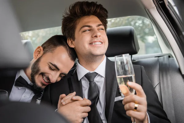 Carefree young gay man in formal wear and braces holding blurred glass of champagne and hand of bearded boyfriend after wedding celebration and sitting on backseat of car — Stock Photo