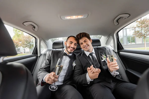 Улыбающаяся гомосексуальная пара в классических костюмах с бутоньерками, держащимися за руки и бокалы шампанского во время празднования свадьбы на заднем сиденье автомобиля — стоковое фото