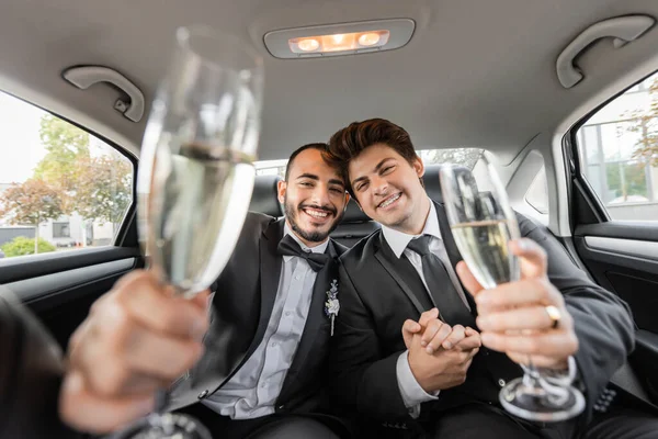 Cheerful homosexual grooms in classic suits holding blurred glasses of champagne and looking at camera during wedding celebration on backseat of car — Stock Photo