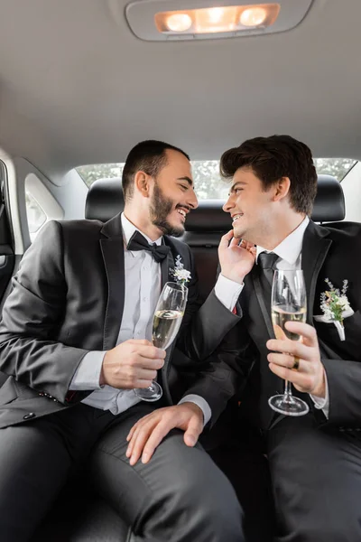 Side view of smiling and bearded gay groom in classic suit touching young boyfriend in braces with glass of champagne while celebrating wedding on backseat of car during honeymoon — Stock Photo