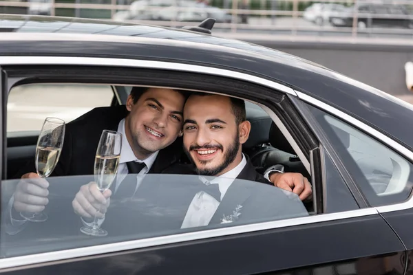 Smiling same sex grooms in formal wear holding glasses of champagne and looking at camera through window while sitting on backseat of car before going to honeymoon — Stock Photo