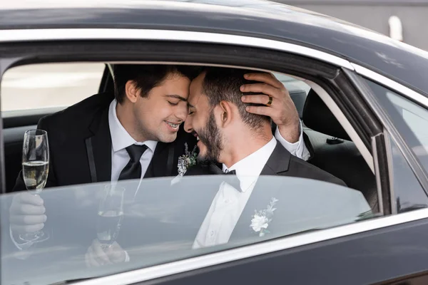 Young homosexual groom in braces and formal wear holding champagne and hugging cheerful bearded boyfriend while while sitting on backseat of car before going to honeymoon — Stock Photo