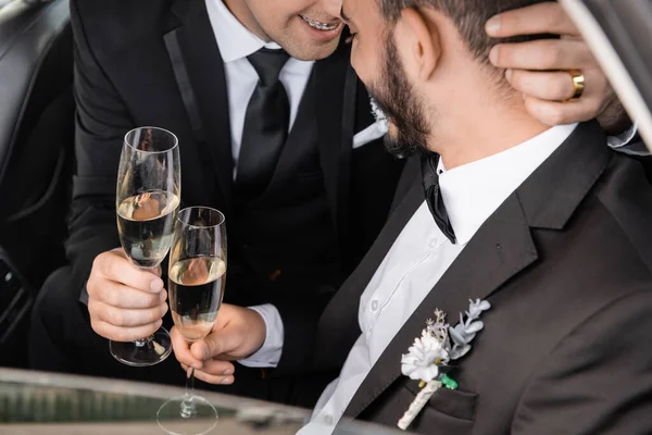 Smiling homosexual groom in formal wear holding champagne glass and hugging bearded boyfriend while celebrating wedding in car during honeymoon — Stock Photo