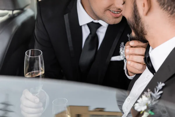 Cropped view of positive gay groom with braces in classic suit touching chin of bearded boyfriend and holding champagne after wedding celebration in car — Stock Photo
