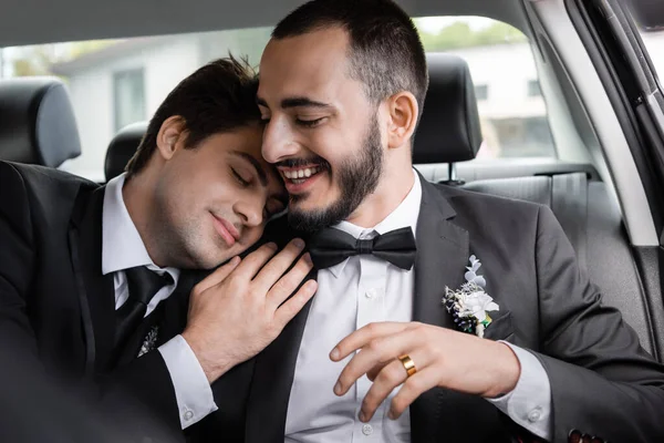 Sonriente hombre barbudo en traje elegante con boutonniere sentado cerca joven novio morena con los ojos cerrados en el coche después de la celebración de la boda mientras va de luna de miel - foto de stock