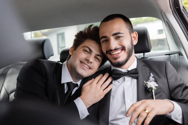 Alegre novio homosexual con tirantes en traje abrazando novio barbudo y mirando a la cámara después de la celebración de la boda mientras va de luna de miel - foto de stock