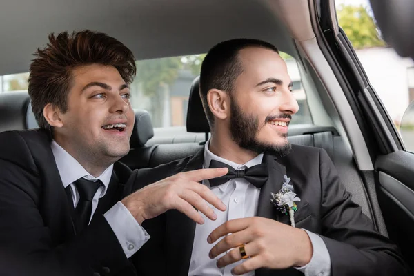 Alegre gay novio en frenos y elegante traje apuntando con dedo cerca barbudo novio y mirando juntos a través de coche ventana después de boda celebración mientras va en luna de miel - foto de stock