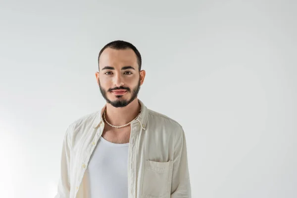 Portrait of young short haired and bearded homosexual man in pearl necklace and casual clothes looking at camera while standing isolated on grey — Stock Photo