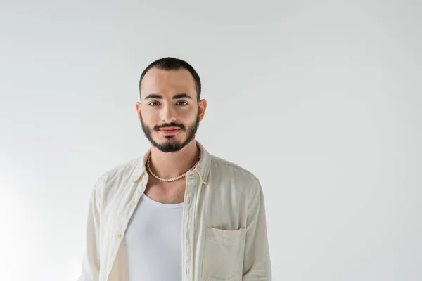 Retrato de joven hombre homosexual de pelo corto y barbudo en collar de perlas y ropa casual mirando a la cámara mientras está de pie aislado en gris - foto de stock