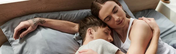 Високий кут зору татуйованої і молодої пари гей в піжамі обережно обіймаються один з одним, сплячи разом на комфортному ліжку вдома вранці, банер — стокове фото