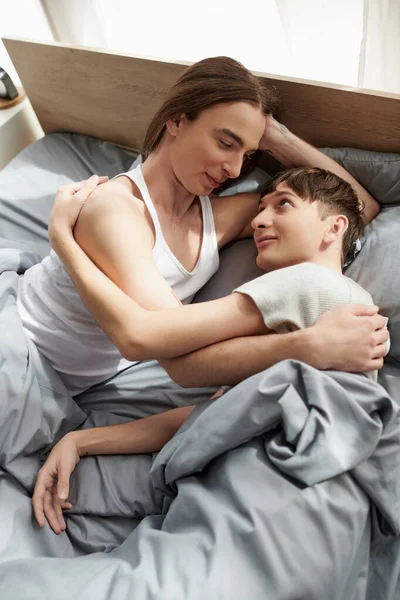 Високий кут зору молодої пари гей в піжамі обіймається і розмовляє один з одним, лежачи разом на зручному ліжку в сучасній спальні вдома вранці — стокове фото