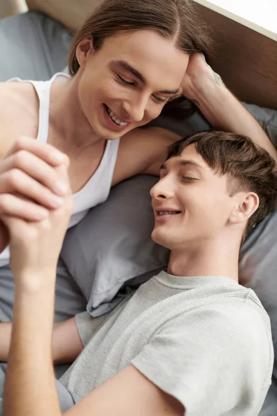 Вид сверху на веселую и счастливую однополую пару в рубашках, держащую друг друга за руки и отдыхающую вместе на удобной кровати дома по утрам — стоковое фото