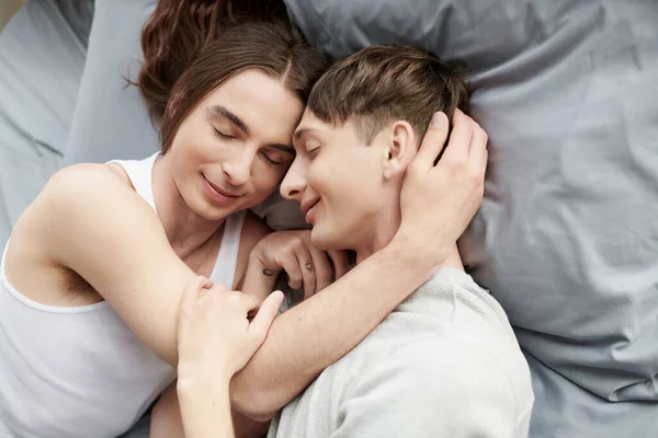 Vue du dessus du couple gay souriant avec les yeux fermés étreignant et se touchant tout en étant couchés ensemble sur un lit confortable et confortable à la maison — Photo de stock