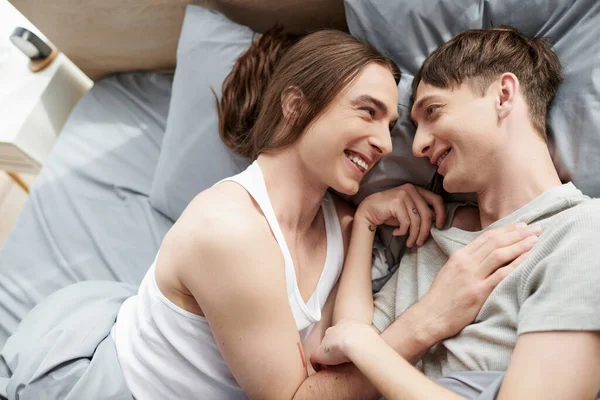 Vue du dessus du couple homosexuel joyeux et tatoué en vêtements de nuit parlant entre eux tout en étant couchés ensemble sur le lit dans la chambre moderne le matin à la maison — Photo de stock