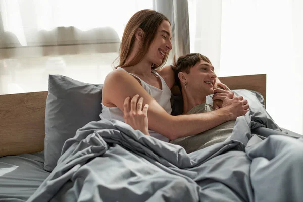 Улыбающийся длинноволосый гей в сонной одежде обнимает парня и смотрит вместе, лежа на удобной кровати в современной спальне утром — стоковое фото