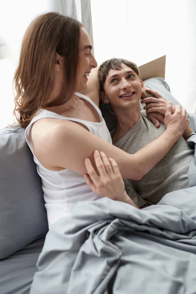 Vista superior de hombre homosexual de pelo largo joven en ropa de dormir abrazo y mirando novio feliz mientras se relajan juntos en la cama en el dormitorio moderno por la mañana - foto de stock