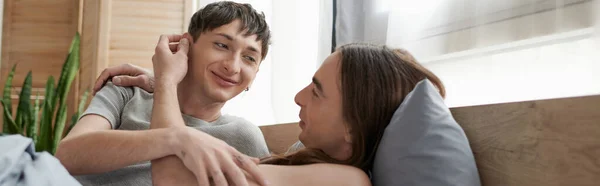 Молодой длинноволосый гей улыбается и трогает ухо татуированного и счастливого партнера во сне, отдыхающего на удобной кровати утром дома, баннер — стоковое фото