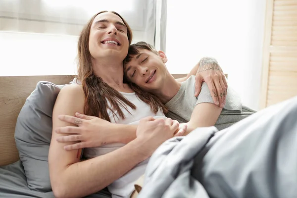 Позитивный и длинноволосый гомосексуалист с закрытыми глазами, обнимающий молодого парня-гея во сне, расслабляясь на современной кровати дома утром — стоковое фото