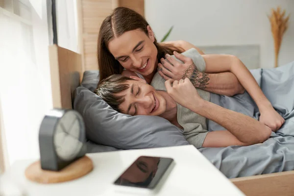 Позитивный и длинноволосый гомосексуалист обнимает молодого парня на кровати рядом с телефоном с пустым экраном и будильником на тумбочке в спальне утром — стоковое фото