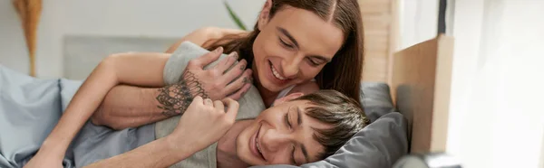 Tatoué et aux cheveux longs gay homme embrassant copain joyeux avec les yeux fermés tout en se réveillant sur un lit confortable le matin, bannière — Photo de stock