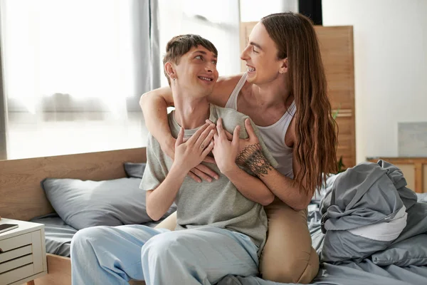Cheerful and long haired gay man with tattoo on hand embracing young boyfriend in pajama while sitting on comfortable bed in bedroom in morning time — Stock Photo