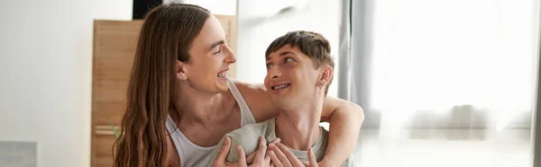 Young long haired gay man in sleepwear hugging carefree boyfriend and looking at each other near curtains with sunlight at modern home in morning, banner — Stock Photo