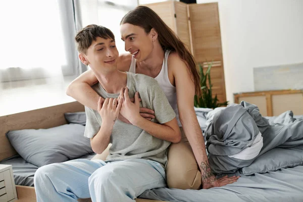 Длинноволосый и татуированный гей обнимается и разговаривает с улыбающимся парнем в пижаме, сидя вместе на кровати в современной спальне в утреннее время — стоковое фото