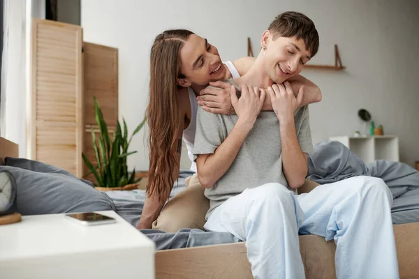 Alegre y de pelo largo gay hombre abrazando y mirando joven novio en pijama en cama cerca de smartphone con pantalla en blanco en mesita de noche en casa por la mañana - foto de stock